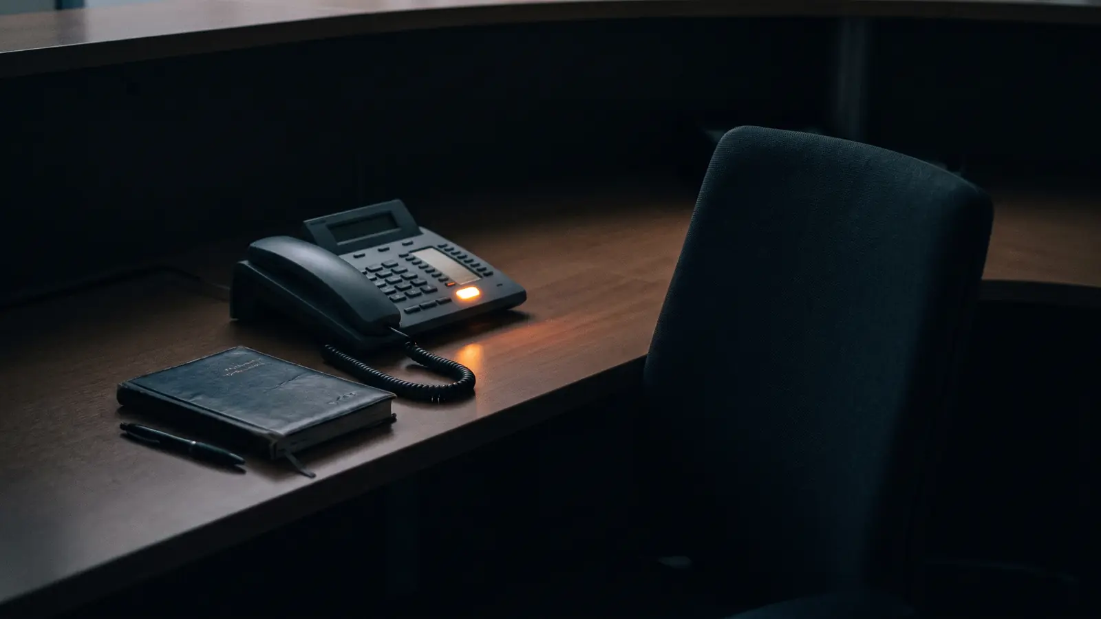 Verlassener Empfangstresen am Abend, auf dem Tisch ein dunkles Tischtelefon, an dem eine einzelne Taste bernsteinfarben leuchtet, daneben ein geschlossener Terminkalender.